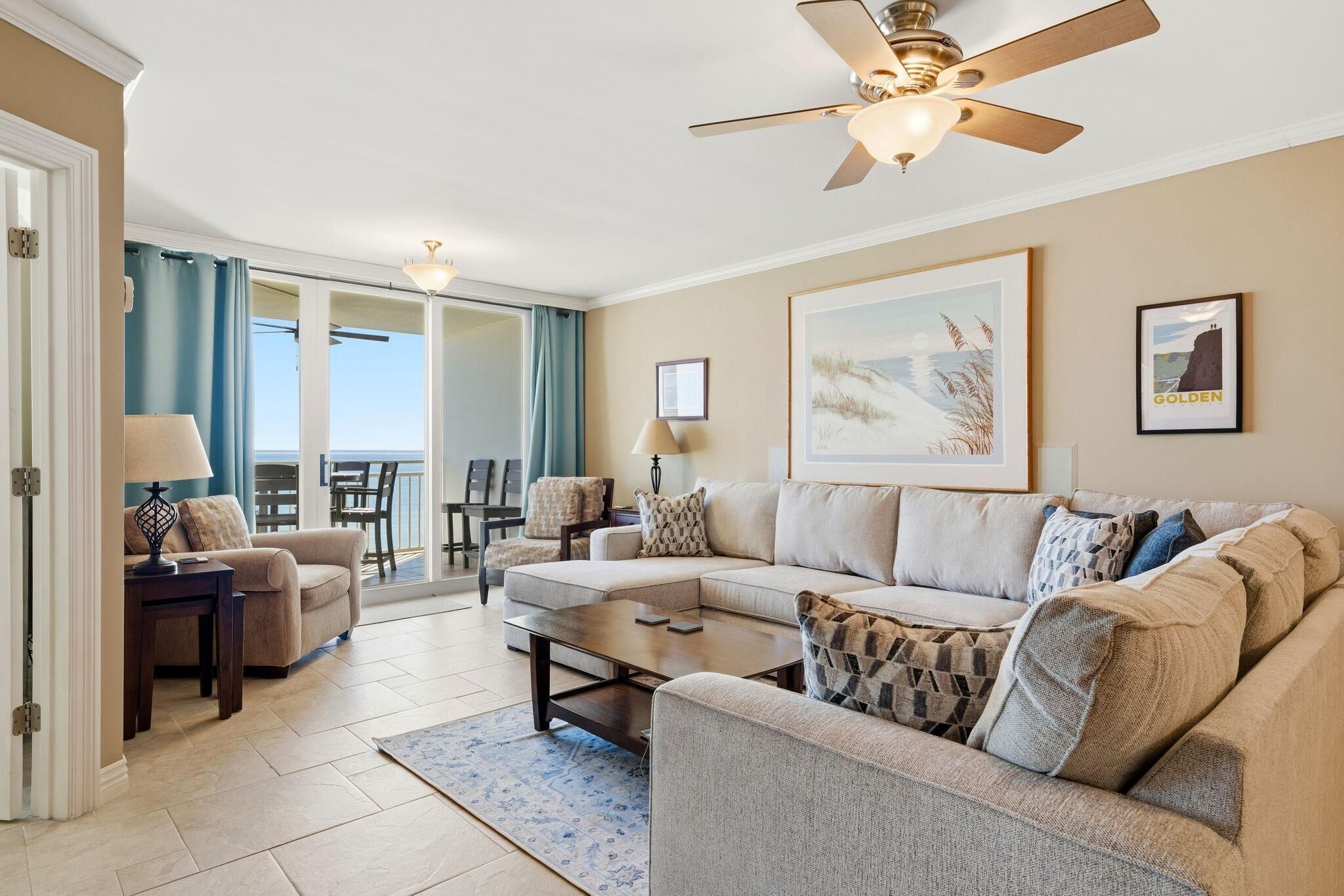 A spacious living room with a sectional sofa, a coffee table, and a ceiling fan. There is a large window with curtains, a framed picture on the wall, and a view of the outdoors.