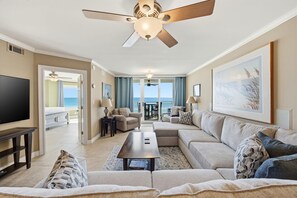 Amplia sala de estar con un sofá modular grande, una mesa de centro y un televisor de pantalla plana. Cuenta con un ventilador de techo, un comedor con mesa y sillas, y vista al océano a través de amplias ventanas.