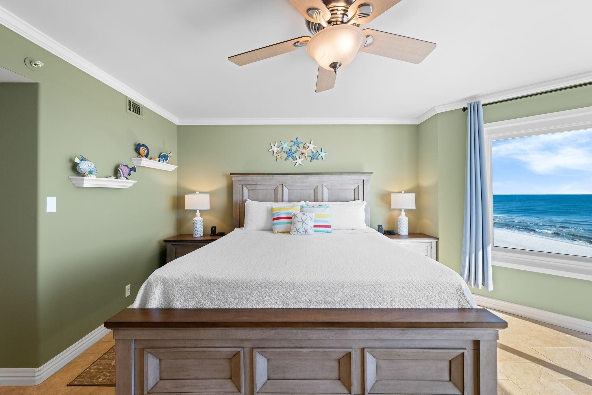 Quarto com cama, mesas de cabeceira, ventoinha no teto, decoração na parede e vista para o mar.