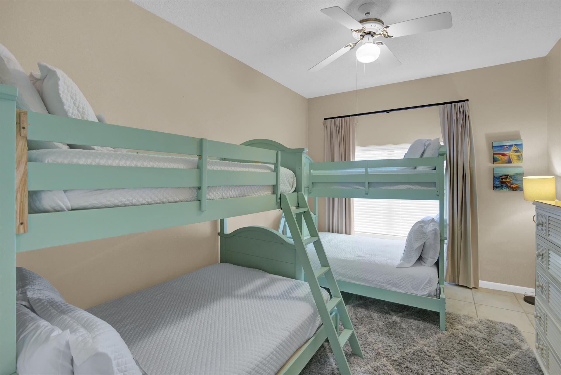 A bunk bed room with a ceiling fan, a window with curtains, and a painting on the wall.