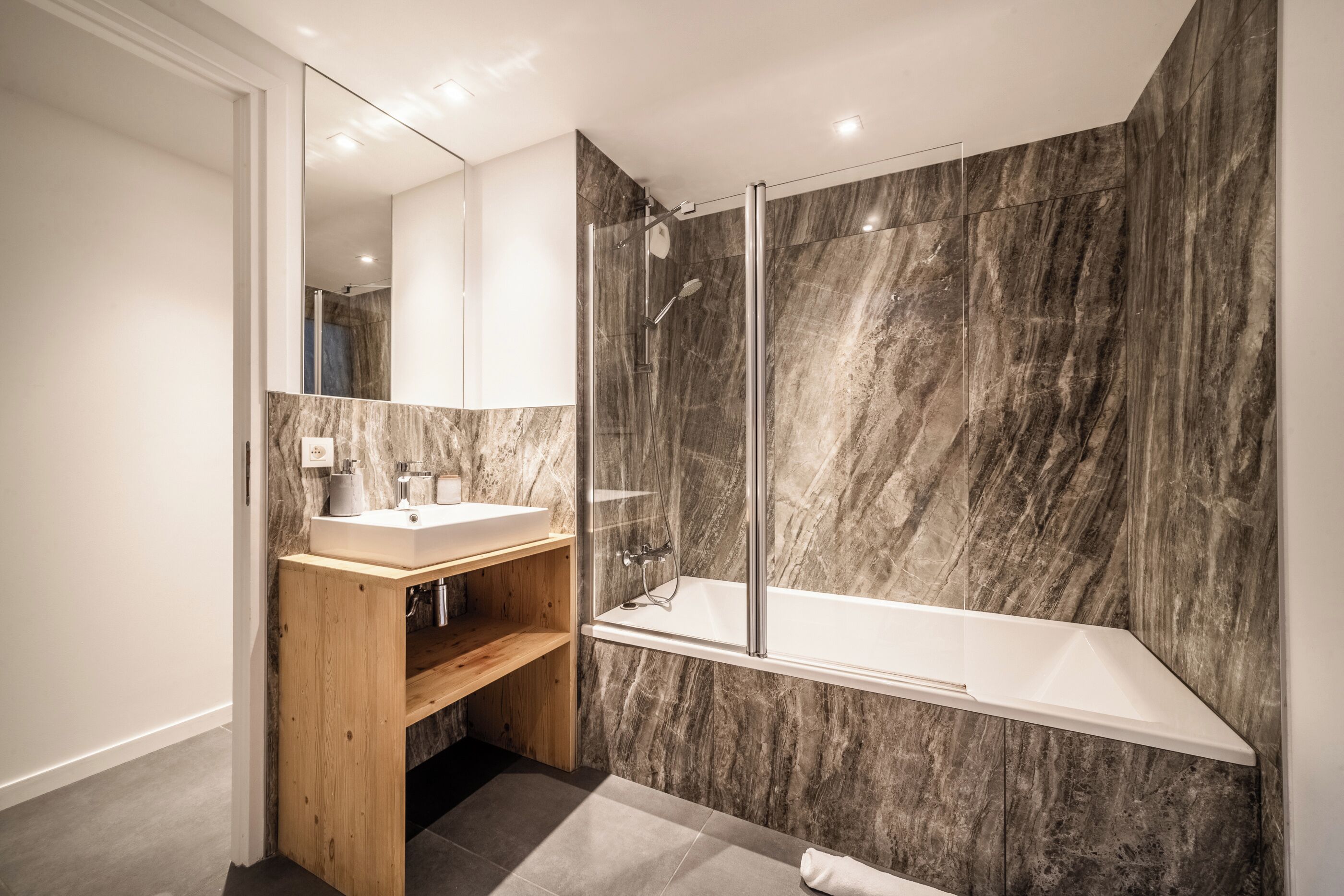 A modern bathroom with a marble wall, a wooden vanity, a glass-enclosed shower, and a white bathtub.