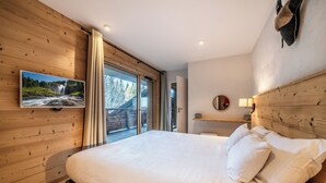 A bedroom with a large bed, wooden walls, a TV mounted on the wall, and a view of a forest through the window.