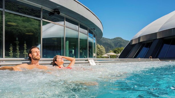 Een stel ontspant in een jacuzzi, met een modern gebouw en bergen op de achtergrond.
