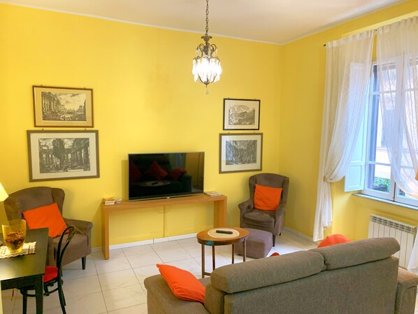 A living room with a television, framed pictures, a chandelier, and a window with curtains. - Rocco's Home at the Colosseum (Roma)