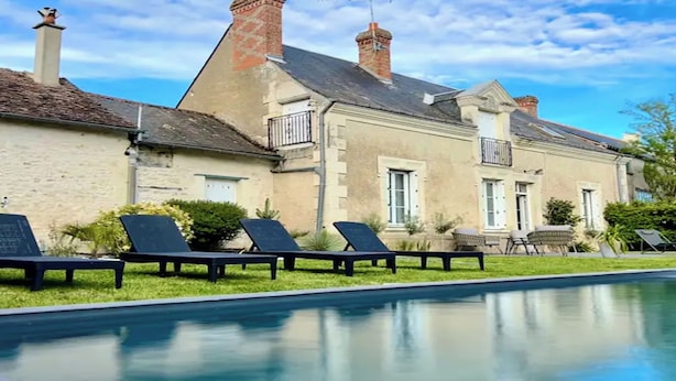 Un espace aménagé au bord de la piscine, avec des chaises longues, une piscine et un bâtiment aux murs de pierre et doté de plusieurs cheminées.