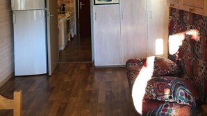 A compact kitchen with a refrigerator, microwave, and cabinets. There is a patterned sofa and a wooden chair.