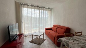 A living room with a red sofa, a TV on a red cabinet, a small table, and a dining table with a patterned tablecloth. - 4th floor, view mountain, balcony, parking, tv, 26m², Villard de Lans (Villard-de-Lans)