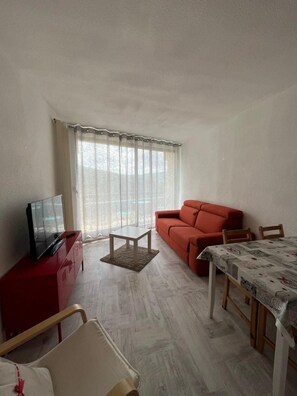 A living room with a red sofa, a TV on a red cabinet, a small table, and a dining table with a patterned tablecloth. - 4th floor, view mountain, balcony, parking, tv, 26m², Villard de Lans (Villard-de-Lans)