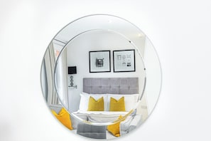 A circular mirror reflecting a bedroom with a grey headboard, white and yellow bedding, and two framed pictures on the wall.
