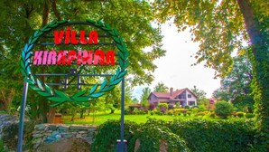 Um letreiro com 'VILLA KIRKIRAN' em letras vermelhas, rodeado por uma grinalda verde, emoldurado por vegetação exuberante e uma casa.