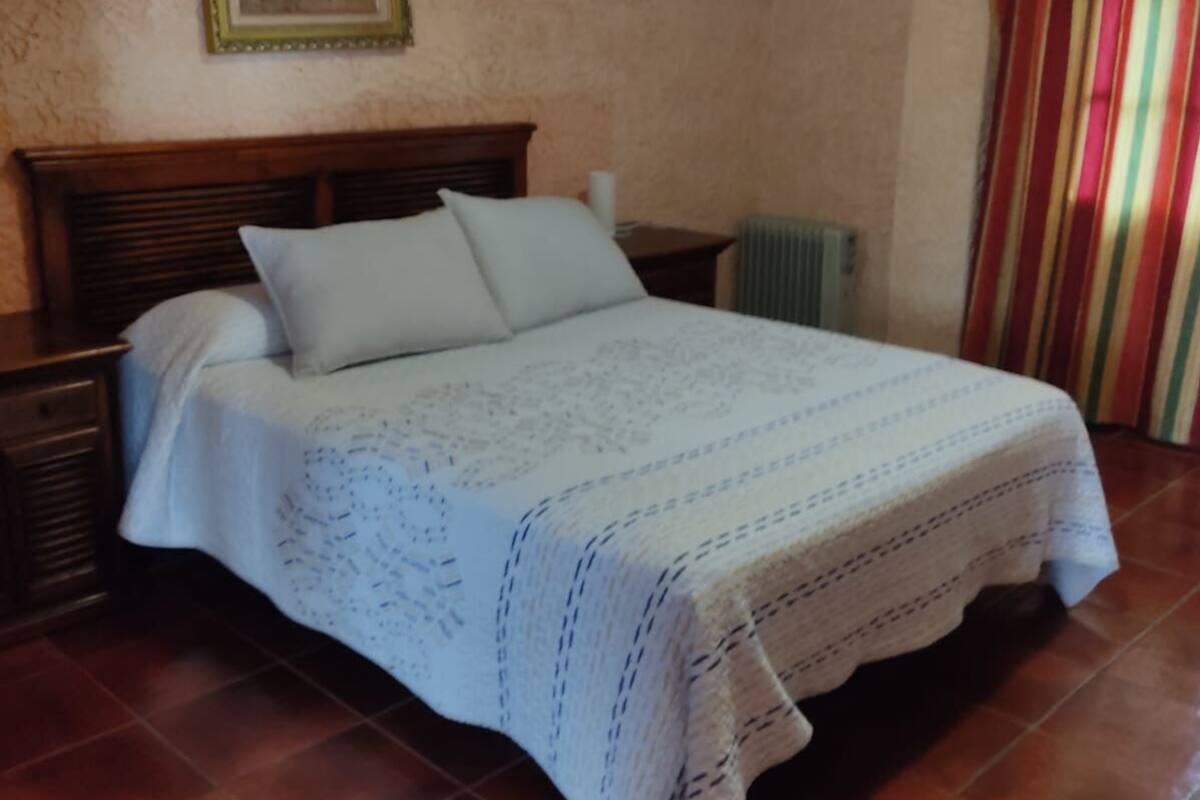 A bedroom with a wooden bed, white bedding, a nightstand, a radiator, and a striped curtain.
