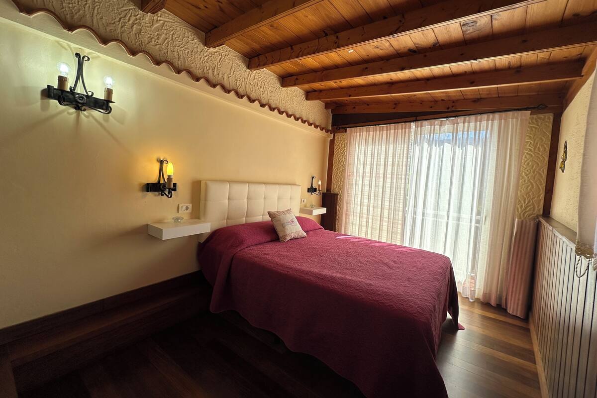 A bedroom with a wooden ceiling, a bed with a red bedspread, a nightstand with a lamp, and a window with sheer curtains.