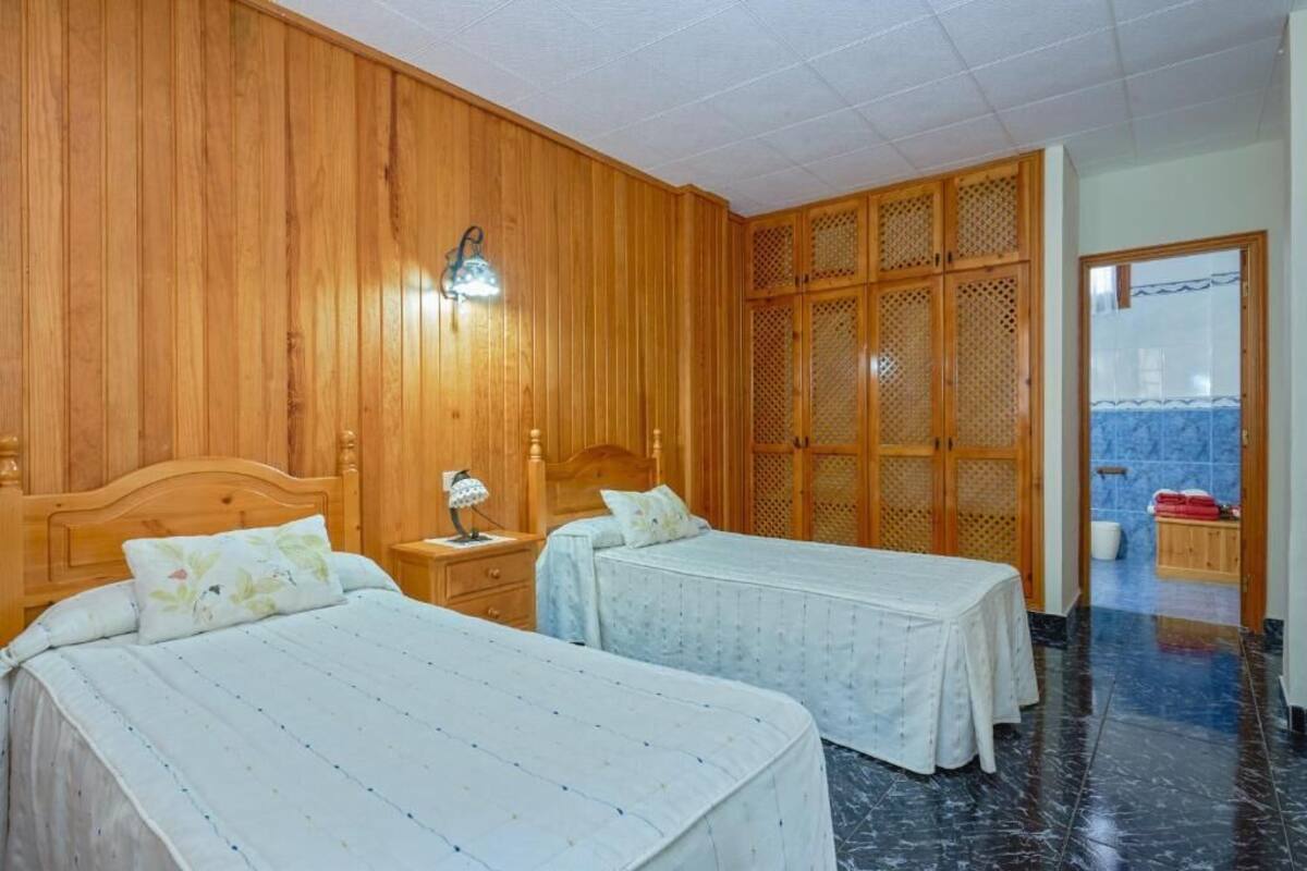 A hotel room with two beds, wooden paneling, a nightstand with a lamp, and a view of the outside through a door.
