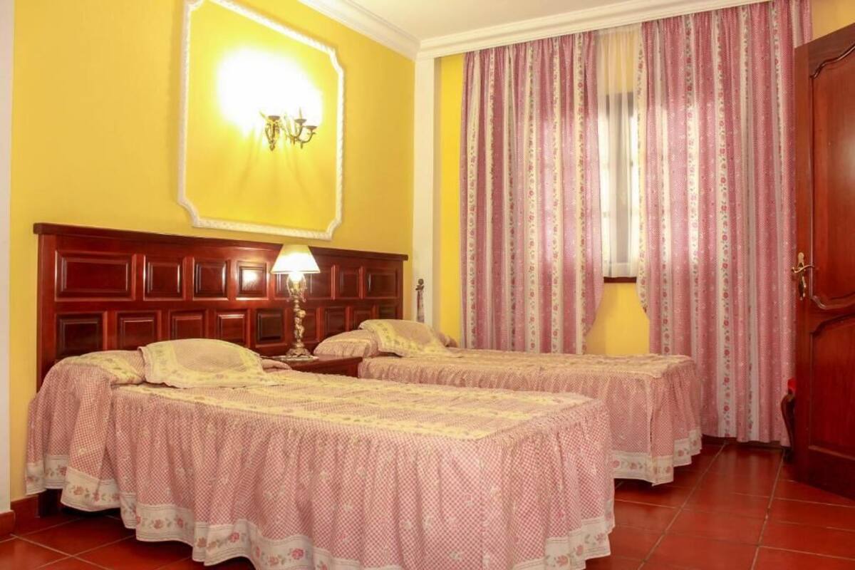 A room with two beds, a wooden headboard, a wall-mounted lamp, and a window with curtains.