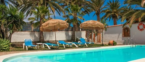 Zona de piscina con sillones reclinables azules y sombrillas de paja, rodeada de palmeras.
