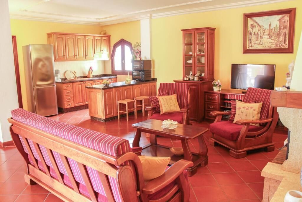 A living room with a sofa, a coffee table, and a TV. There is a kitchen area with wooden cabinets and a refrigerator.