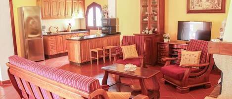 A living room with a sofa, a coffee table, and a TV. There is a kitchen area with wooden cabinets and a refrigerator.