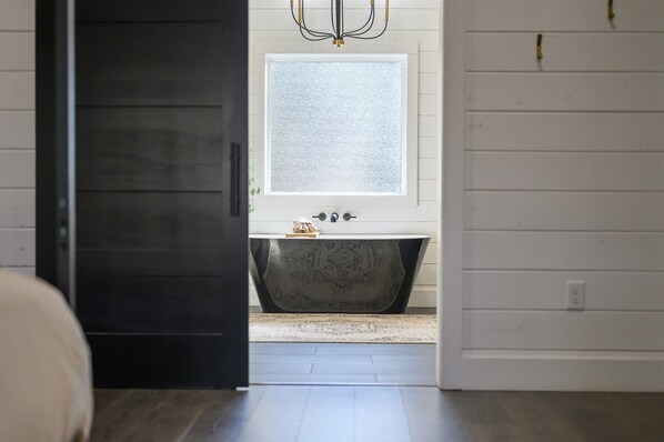 A modern bathroom with a freestanding bathtub, a window above the sink, and a dark door leading to another room. - Romantic,Spacious,Modern Cabin | Hot Tub! (Broken Bow)
