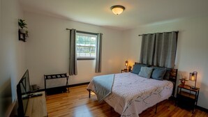 Une chambre à coucher avec un lit, une table de chevet avec une lampe, une fenêtre avec des rideaux et une petite étagère avec des plantes.