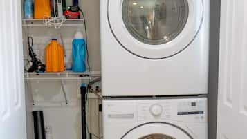 Un cuarto de lavado con dos lavadoras apiladas, un estante con productos de limpieza, un aspirador y una cesta de ropa.
