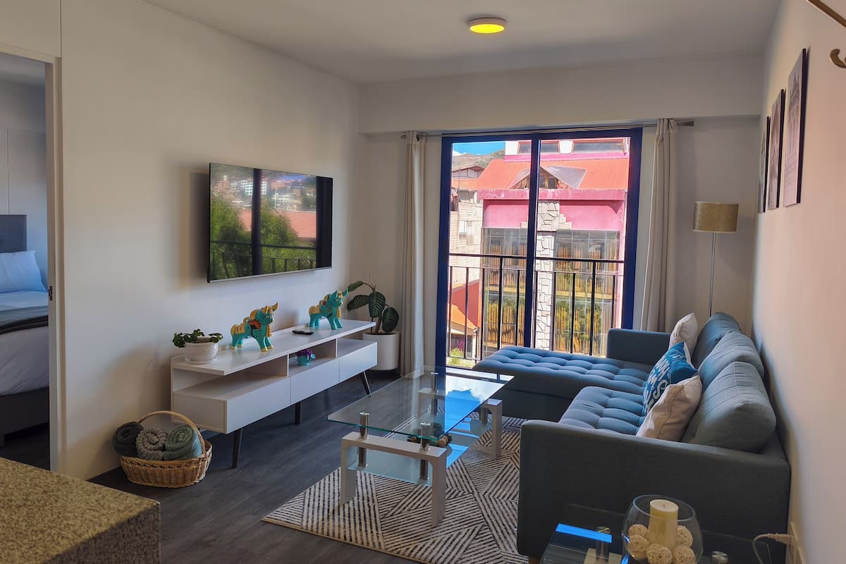 Una sala moderna con sofá, una mesa de centro de vidrio, televisor montado en la pared, balcón con vistas a edificios y un mueble blanco para entretenimiento con objetos decorativos.