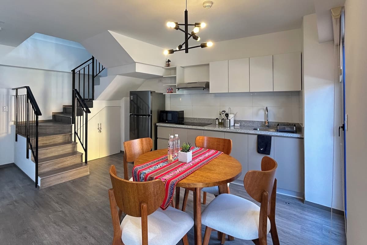 Cocina moderna con armarios blancos, microondas integrado y refrigerador de acero inoxidable. Hay un comedor con mesa y sillas de madera, y una escalera que sube a un nivel superior.