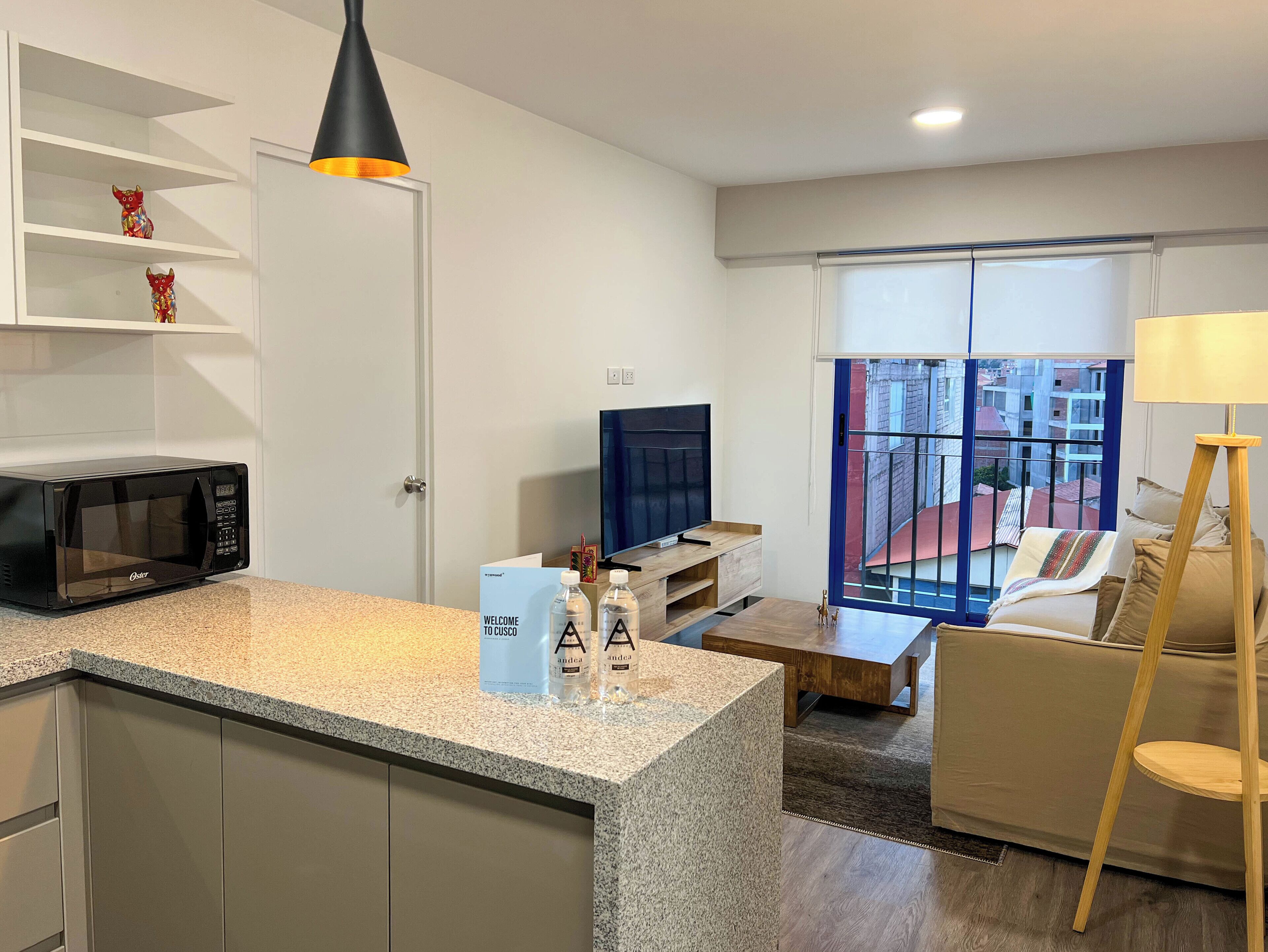 A modern kitchen with a built-in microwave, a coffee machine, and a small refrigerator. There is a dining area with a wooden table and chairs, and a sofa with a view of the city.