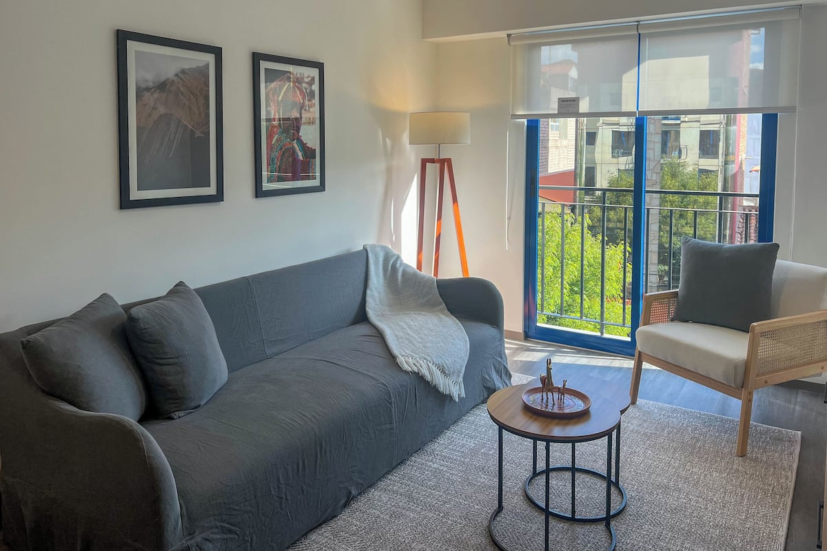 Una sala moderna con un sofá gris, una mesita redonda pequeña y un sillón. Hay dos cuadros enmarcados en la pared, una lámpara de pie y un amplio ventanal con vistas a zonas verdes y edificios.