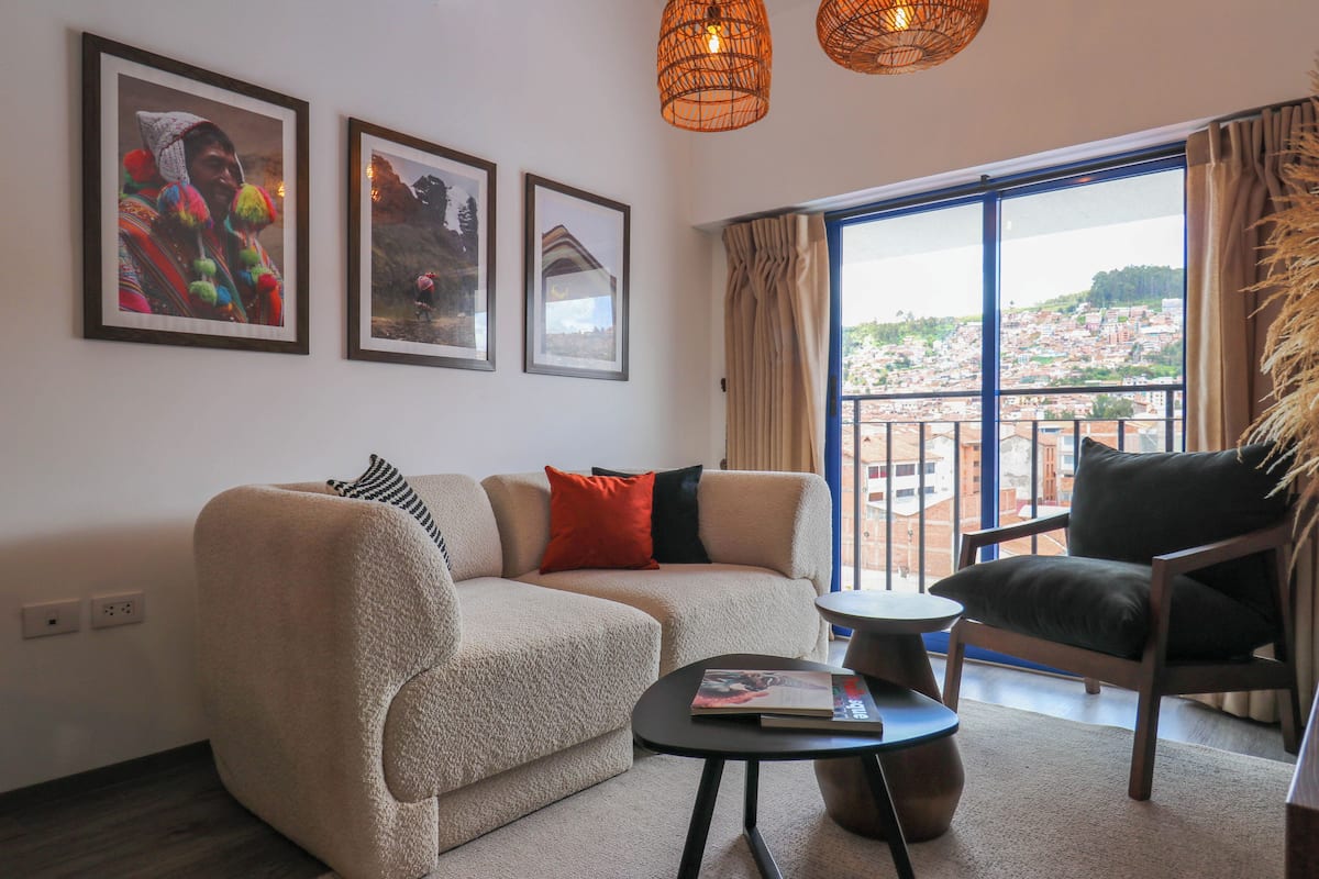 Una sala de estar con sofá, sillón y una mesa de centro. En la pared, hay cuadros enmarcados y vistas de un paisaje urbano a través de amplias ventanas.