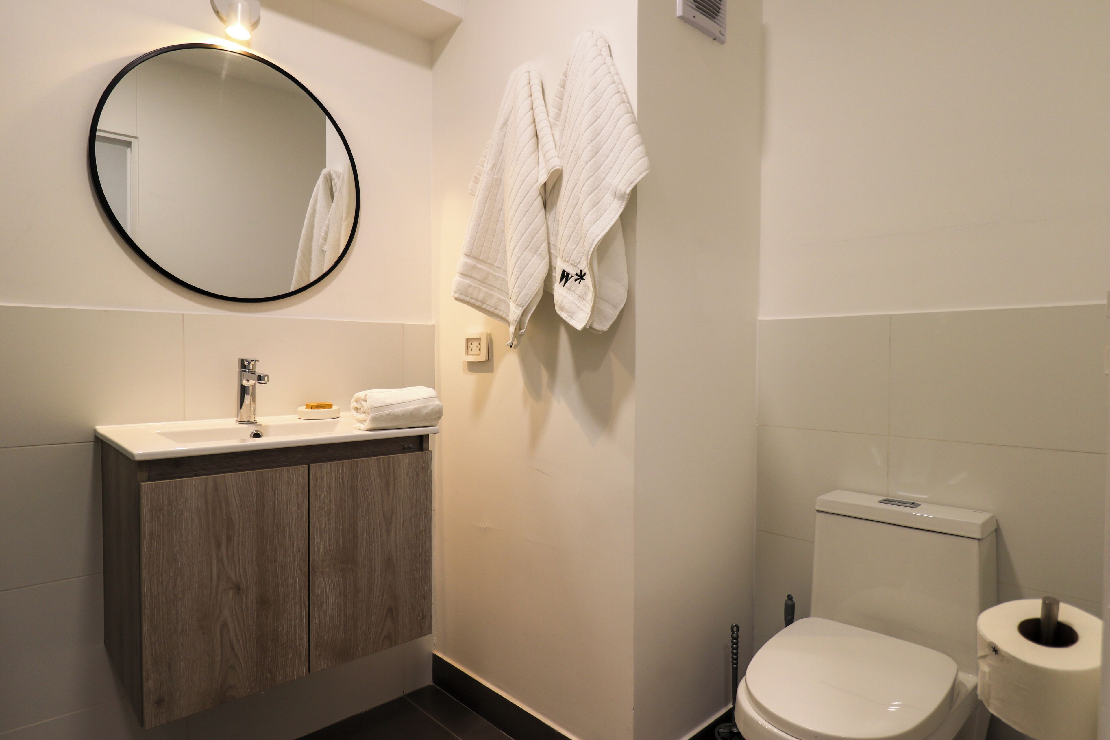 A bathroom with a round mirror, a sink with a wooden cabinet, a toilet, and two white towels hanging on the wall.