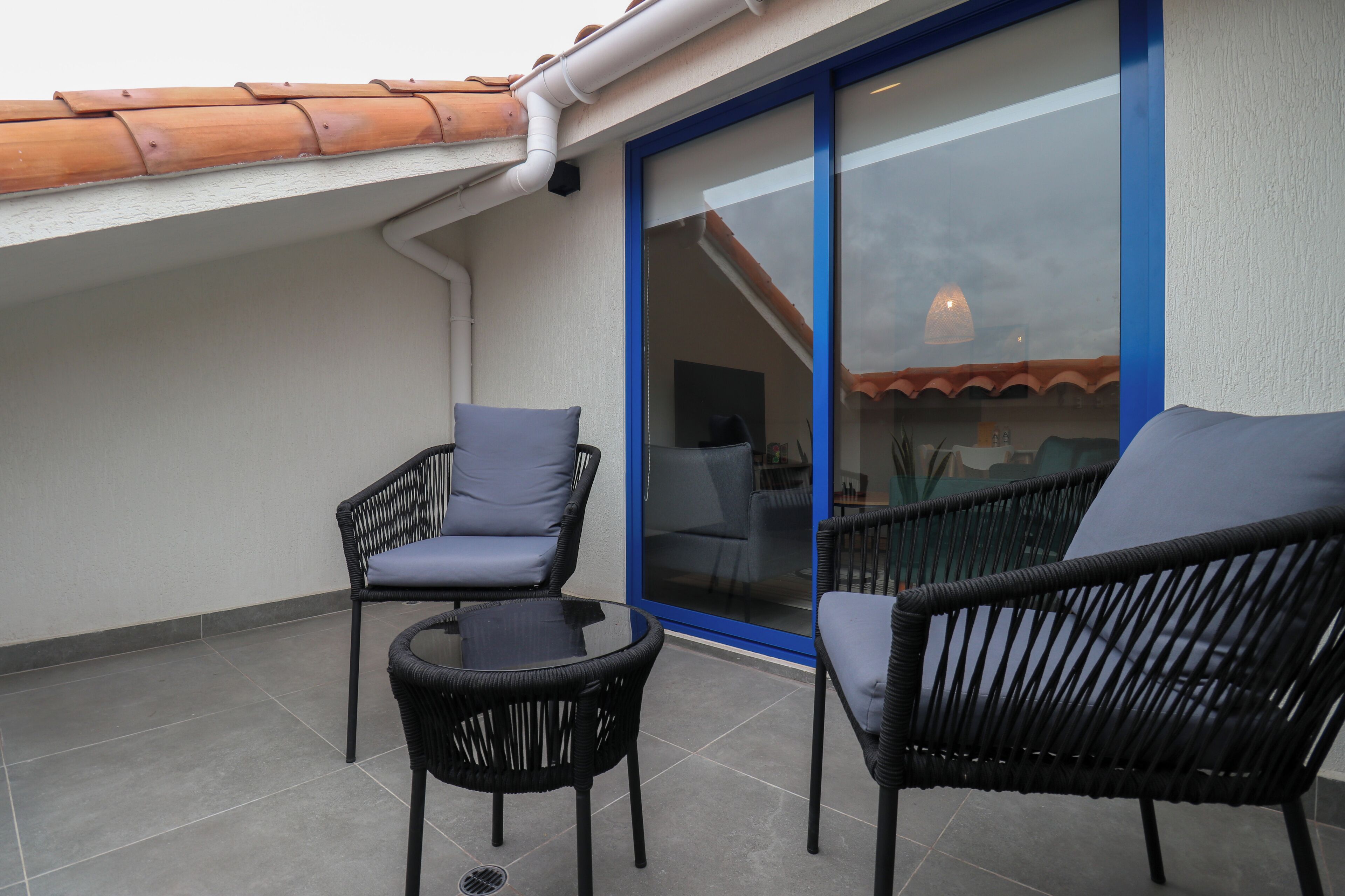 A balcony with two wicker chairs and a small table, a sliding glass door, and a tiled floor.