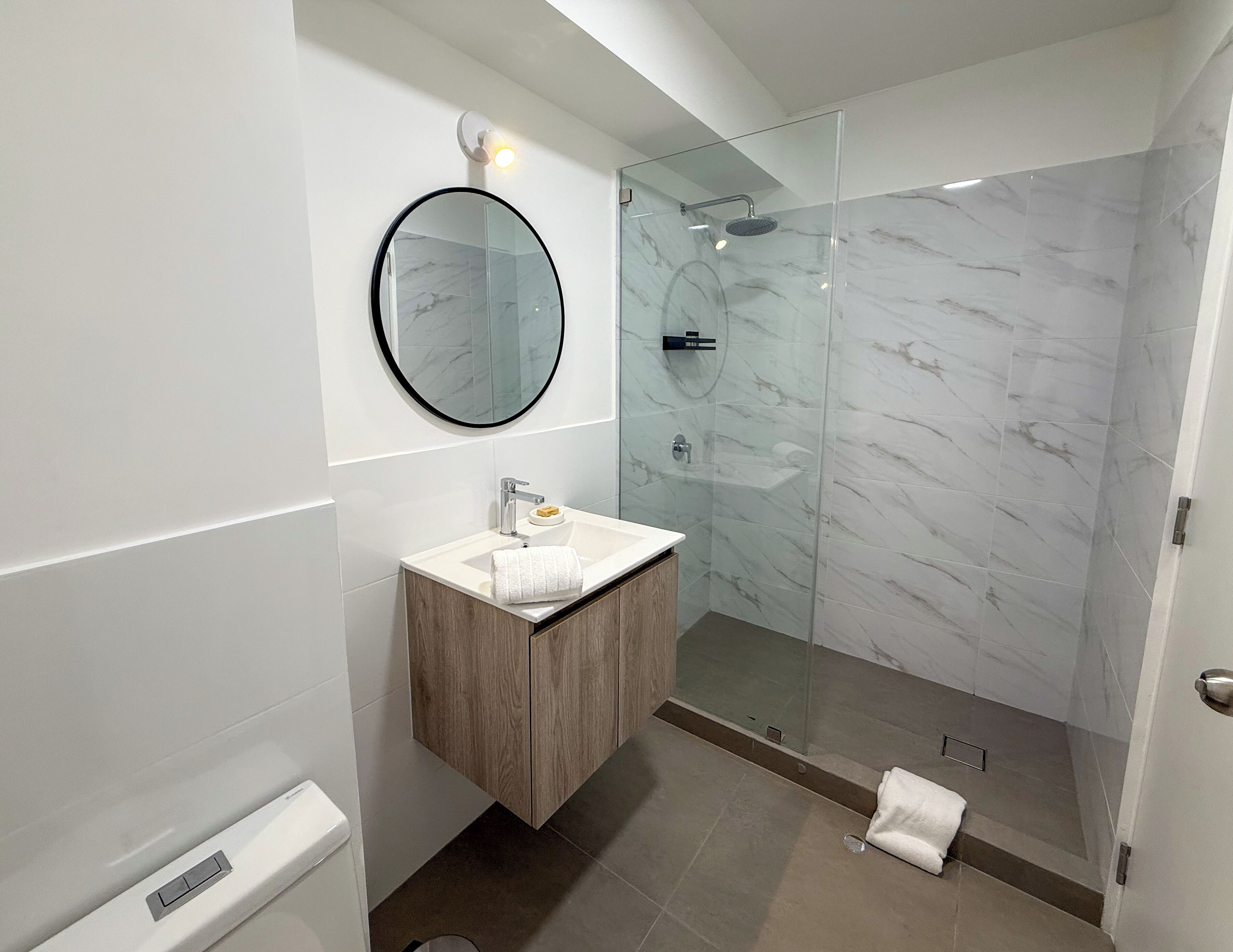 Un baño moderno con ducha de mármol, espejo redondo, tocador de madera y lavabo blanco.