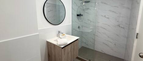 A modern bathroom with a marble shower, a round mirror, a wooden vanity, and a white sink.