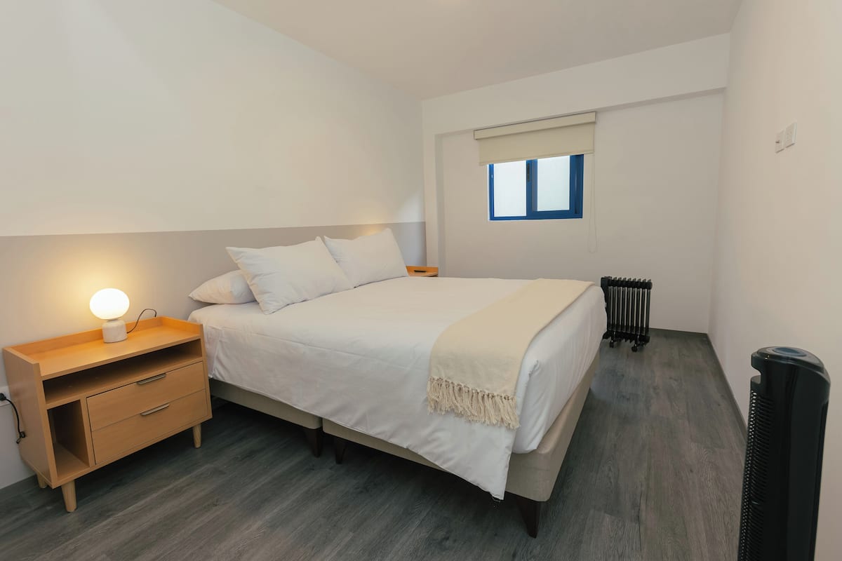 Un dormitorio con cama, mesita de noche, lámpara, ventana, radiador y cubo de basura.