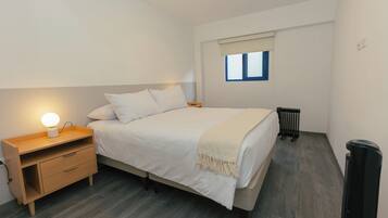 A bedroom with a bed, bedside table, lamp, window, radiator, and a trash bin.