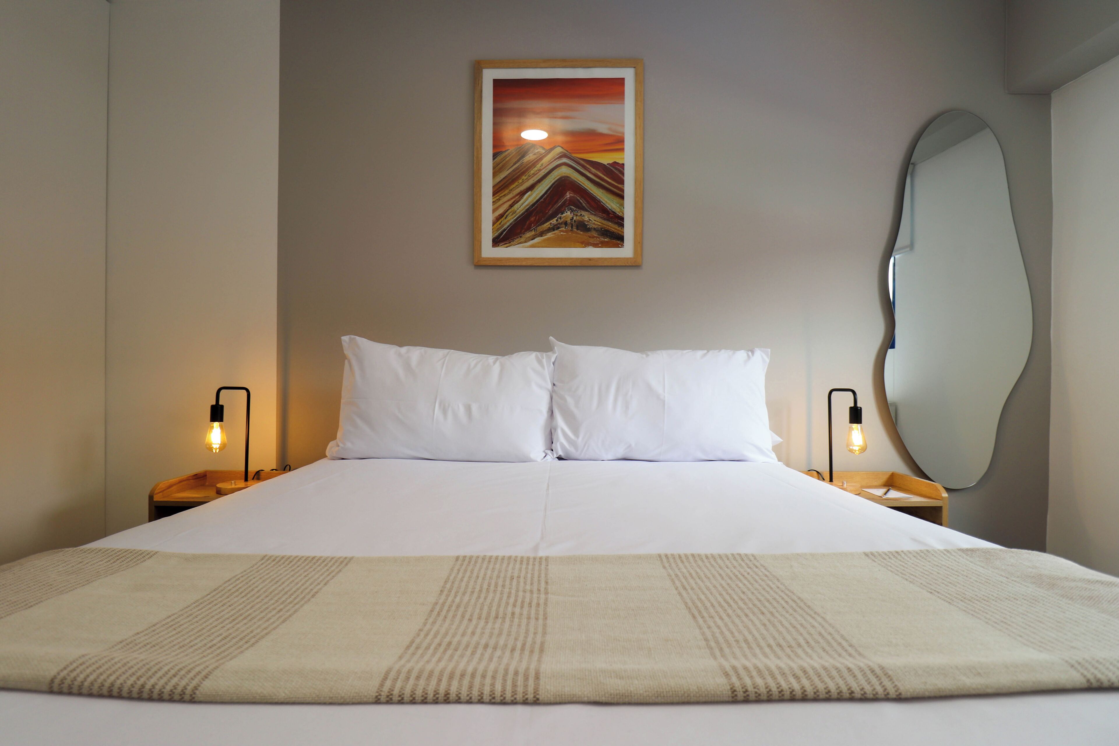 Una cama bien hecha con ropa blanca, dos lámparas de noche y un cuadro de un atardecer sobre montañas en la pared.