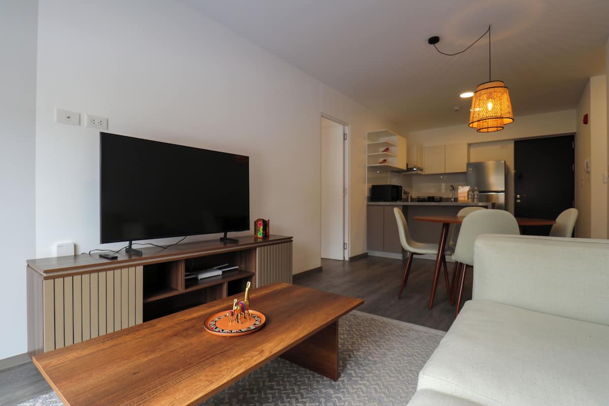 Una sala de estar moderna con un televisor de pantalla plana, una mesa de centro de madera y un sofá. Se observa una zona de cocina con un refrigerador y microondas al fondo.