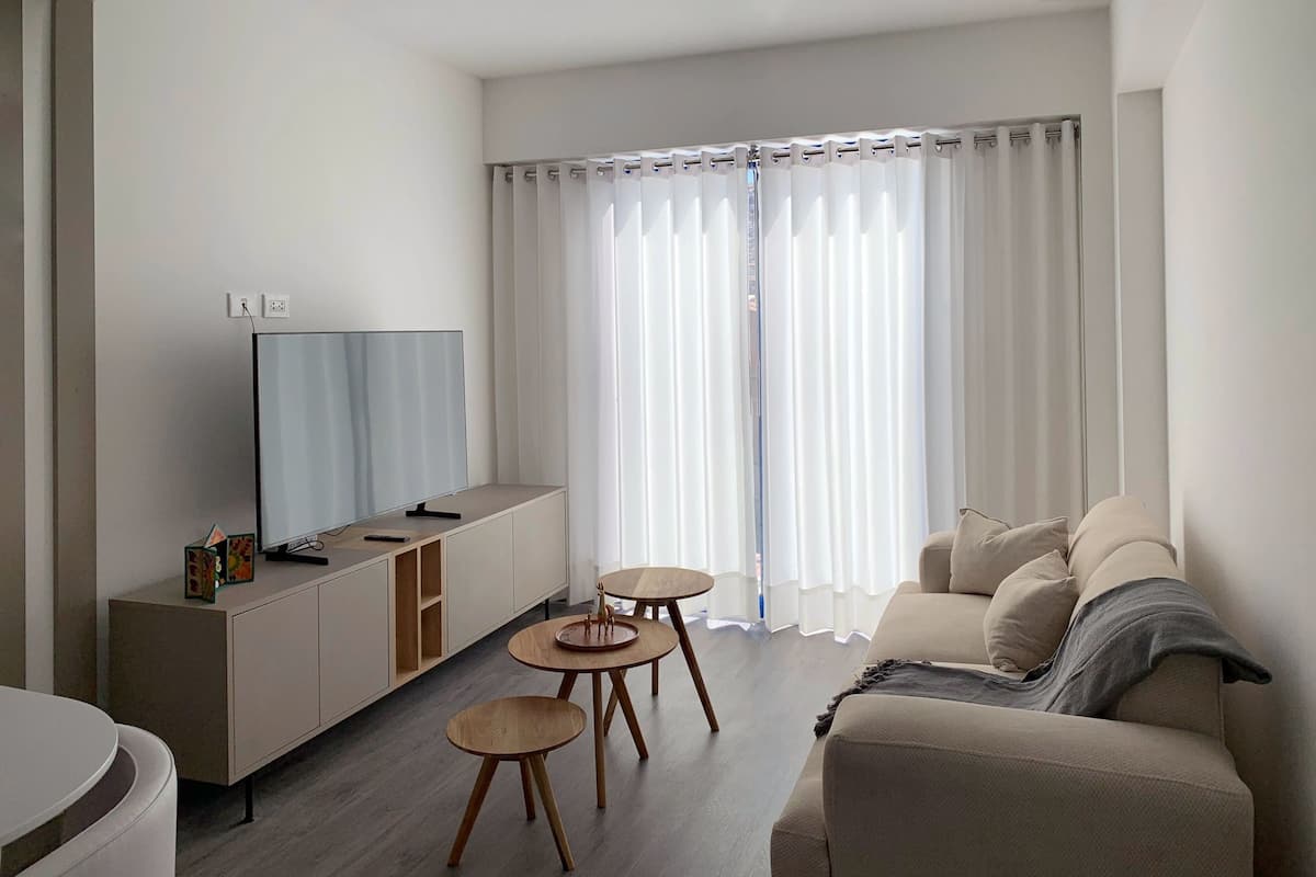 Una sala de estar moderna con un televisor de pantalla plana, un sofá, dos mesitas de madera y un gran ventanal con cortinas de gasa.