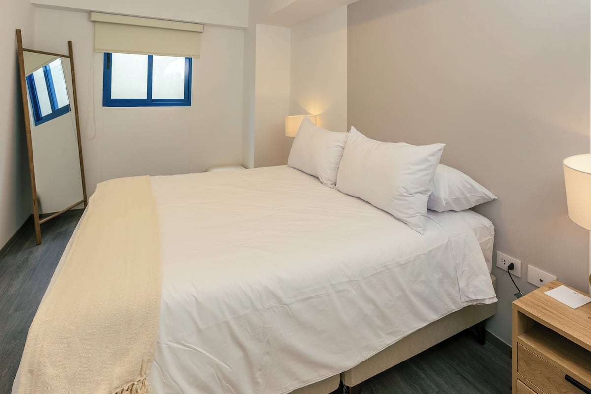 Una cama bien hecha con sábanas blancas, una mesita de noche con lámpara y una ventana con marco azul.