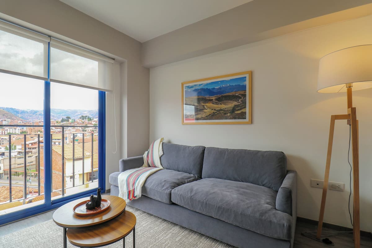 Un salón con un sofá gris, una mesita con un objeto decorativo, una lámpara de pie y un ventanal que ofrece vistas a la ciudad.