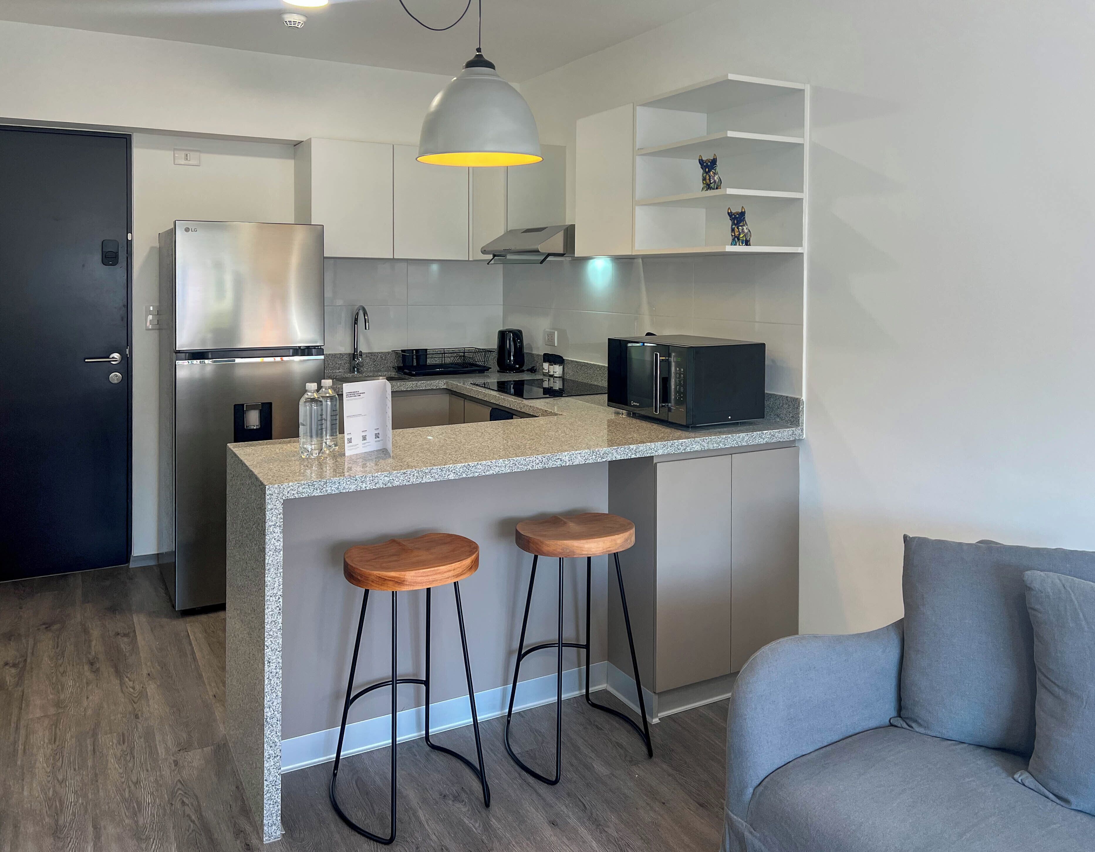 A modern kitchen with a stainless steel refrigerator, a microwave, and a coffee maker. There are two wooden stools and a grey sofa in the living area.
