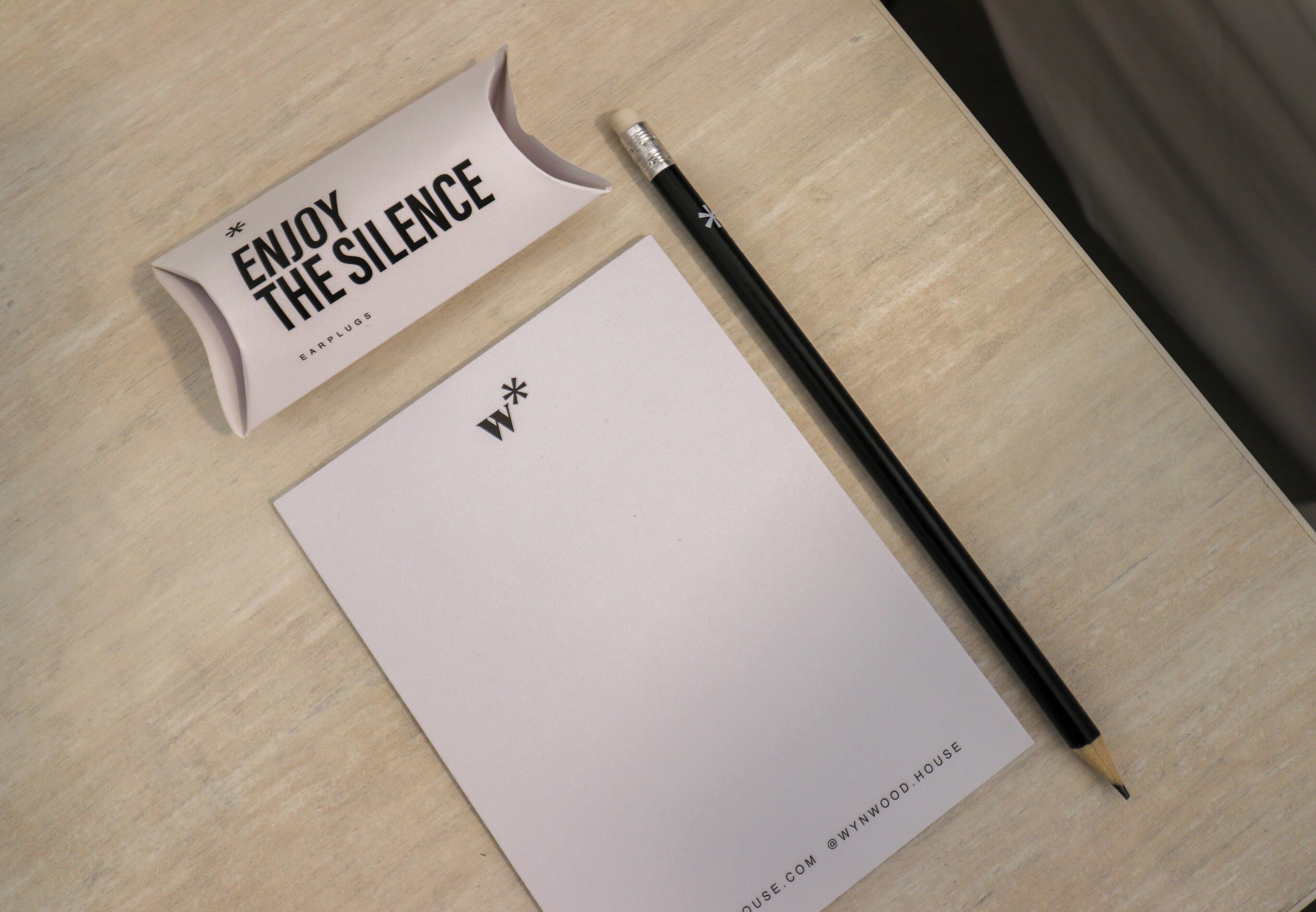 A wooden desk with a white notepad, a black pencil, and a pink box labeled "ENJOY THE SILENCE EARPLUGS."