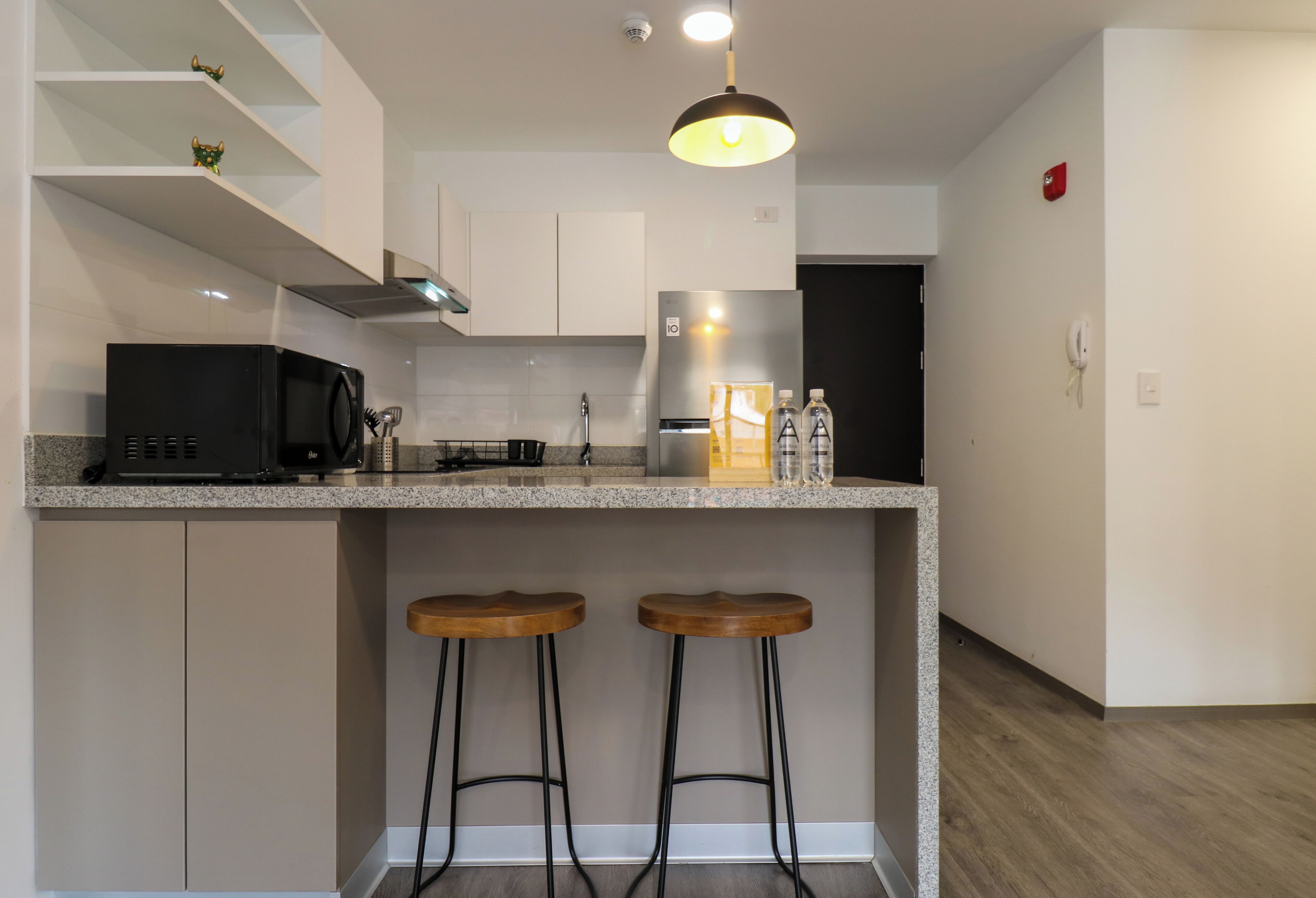 A modern kitchen with a built-in island, bar stools, a microwave, a refrigerator, and a pendant light.