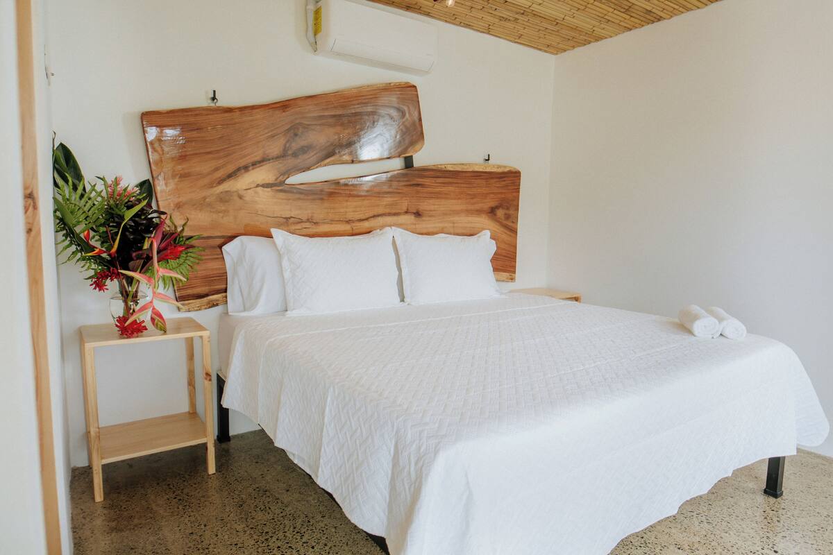 A bedroom with a wooden headboard, a white bedspread, and a small wooden nightstand with a vase of flowers.