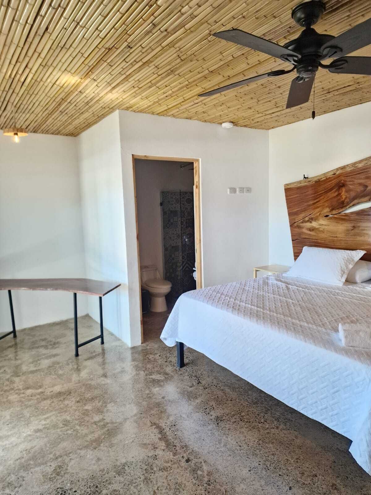 Un dormitorio con cama, escritorio, ventilador de techo y baño visible a través de una puerta abierta.