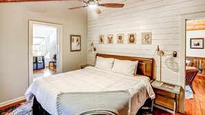 A bedroom with a wooden ceiling, a bed with white bedding, a bedside table with a lamp, and framed pictures on the wall.