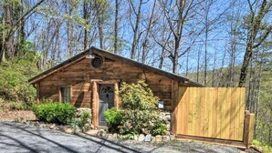 A wooden cabin with a porch, surrounded by trees and a gravel driveway.