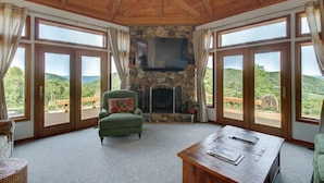 A cozy room with a stone fireplace, a green armchair, and a wooden table. Large windows offer a view of the surrounding landscape.