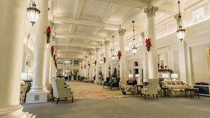 A spacious lobby with high ceilings, ornate columns, and chandeliers. There are multiple seating areas with chairs and sofas, and a carpeted floor.