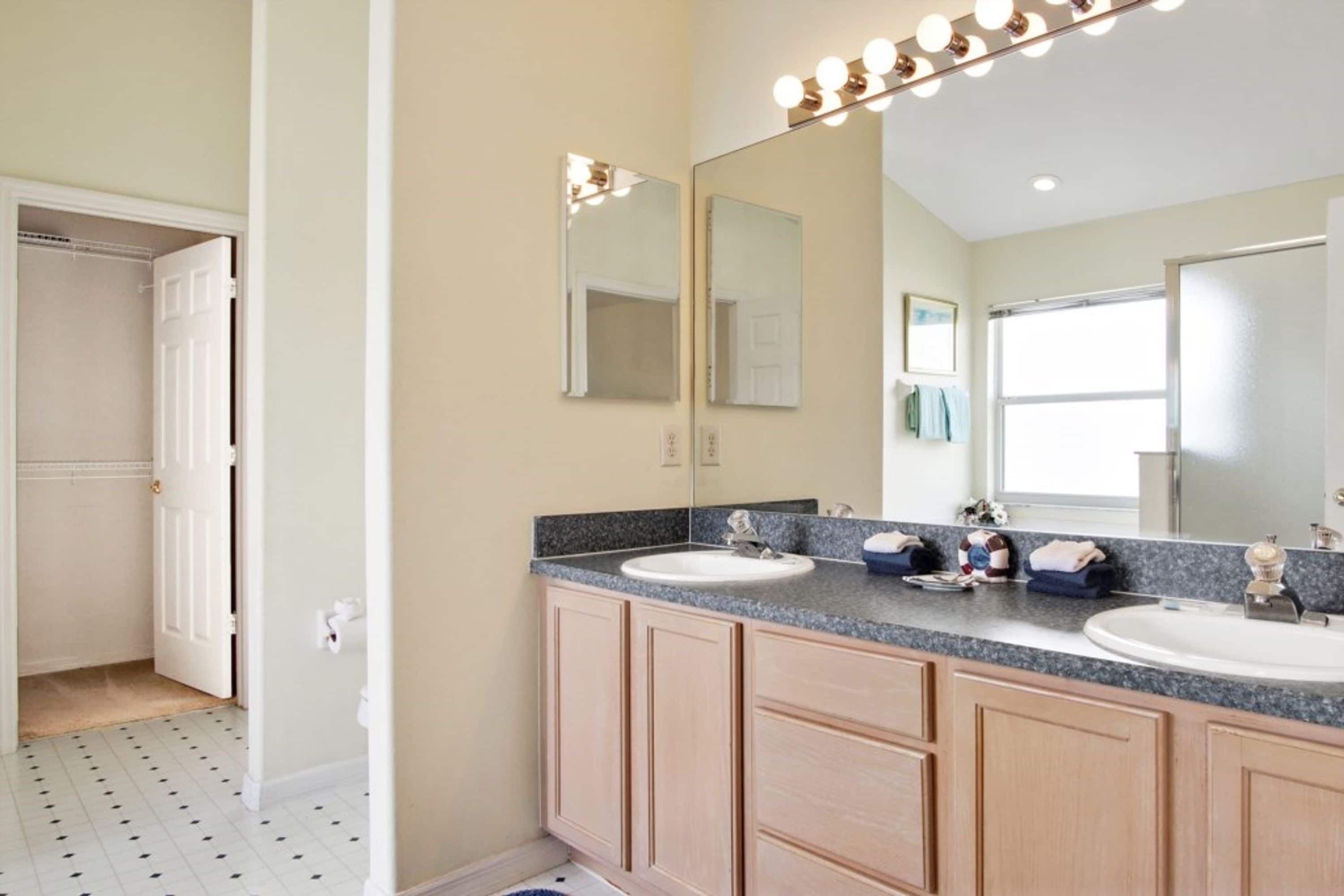 A bathroom with two sinks, a large mirror with lights, wooden cabinets, and a tiled floor.